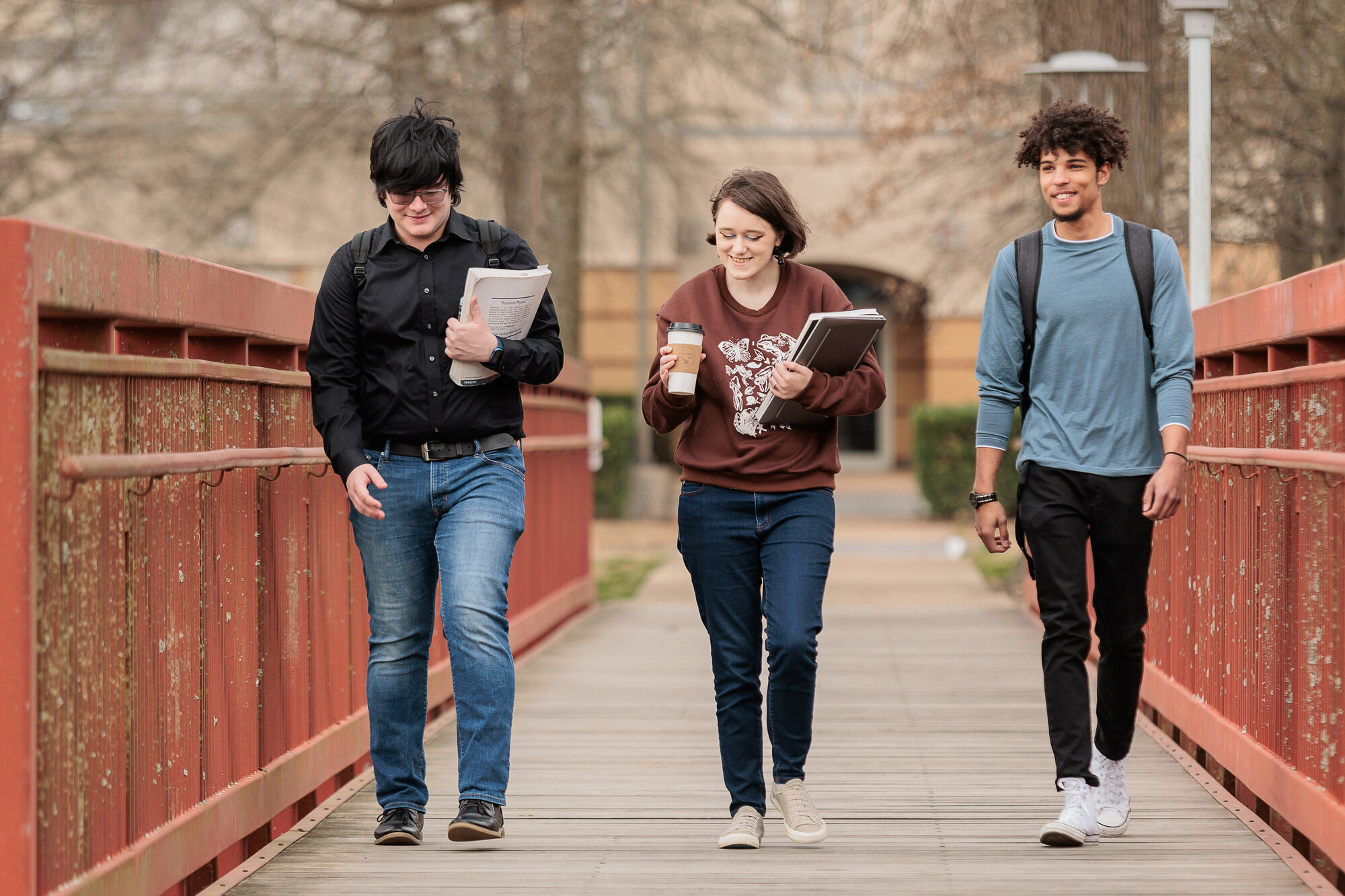 Three college students walk together across a red pedestrian bridge on campus. The student on the left wears a black shirt and jeans, holding papers and a backpack. The student in the middle wears a maroon sweatshirt with a white design, carrying a laptop and coffee cup. The student on the right wears a blue long-sleeve shirt, black pants, and a backpack. They are smiling and engaged in conversation, with a blurred academic building and trees in the background.