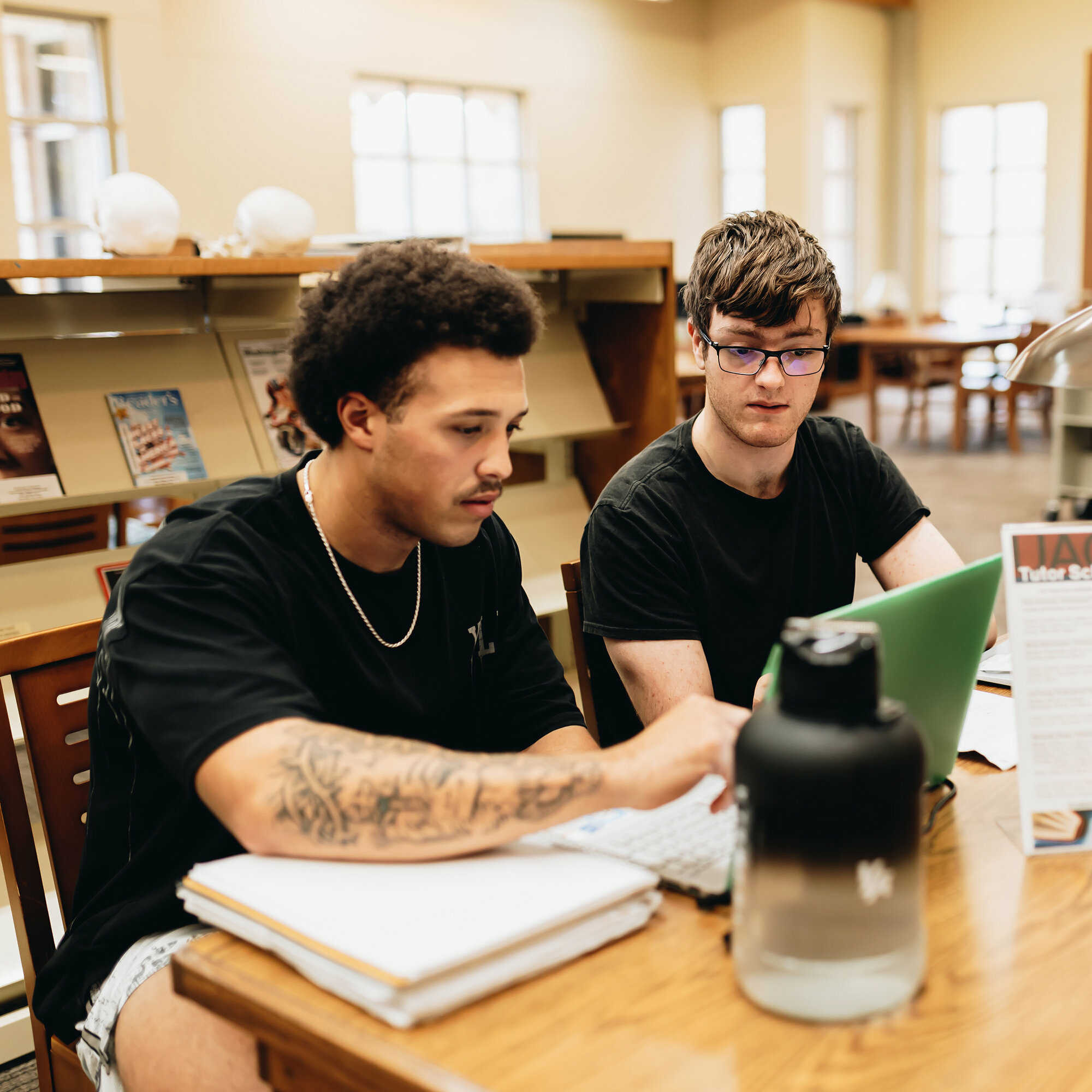 Two college students work together at a library table. One student points at a laptop screen while the other listens attentively. Books, notebooks, and a large water bottle sit on the table. The background features shelves, magazines, and large windows letting in natural light.