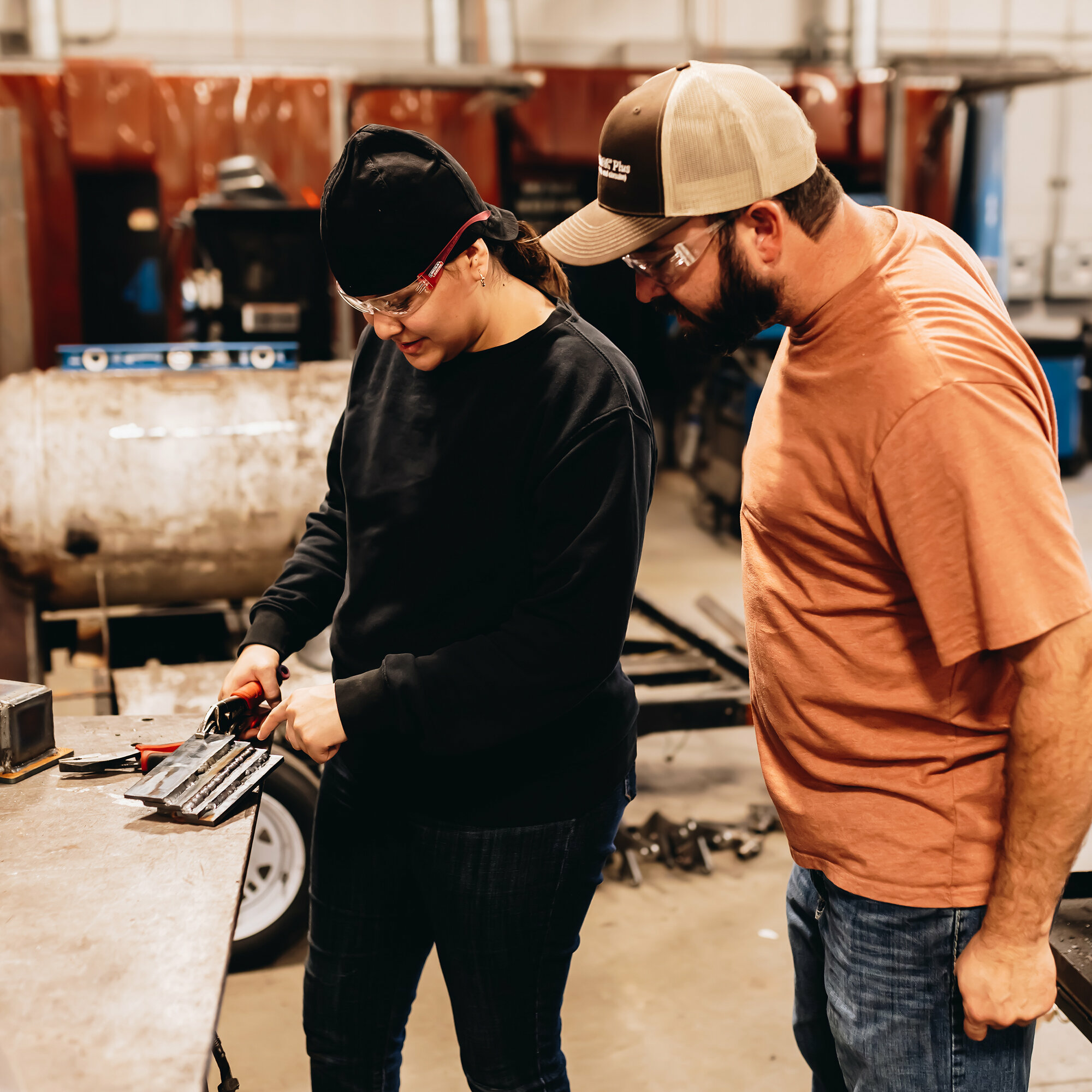 A welding instructor and student work together in a workshop. The student, wearing safety glasses, a welding cap, and a black sweatshirt, uses a tool on a metal piece while the instructor, in a tan cap, safety glasses, and orange shirt, observes closely and provides guidance. The background shows welding equipment and workshop tools.