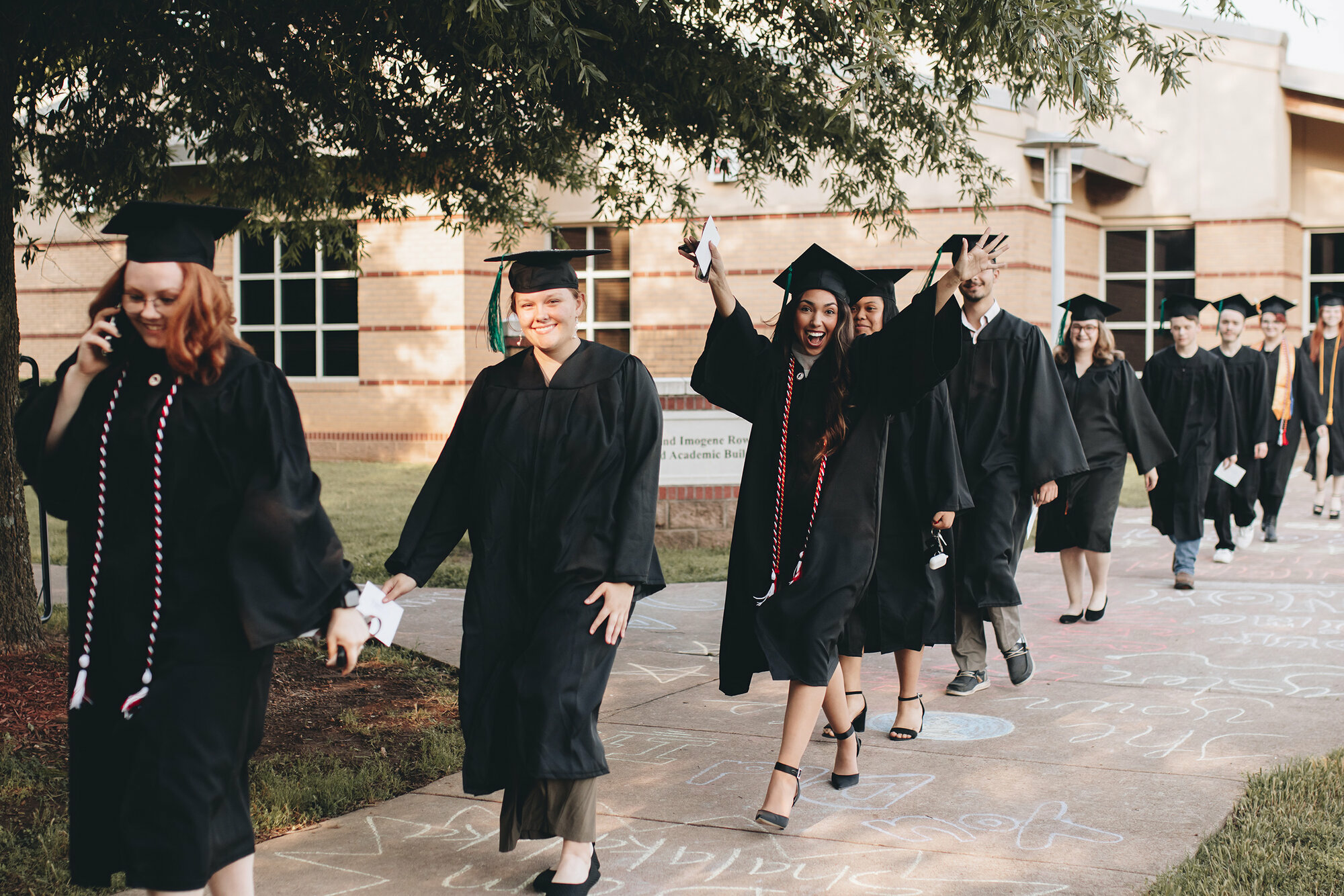 A line of graduates in black caps and gowns walks along a sidewalk outside a campus building during commencement. Some students smile, wave, and celebrate while holding their diplomas, and others chat or take photos. The path is decorated with colorful chalk messages, and trees provide shade along the route.