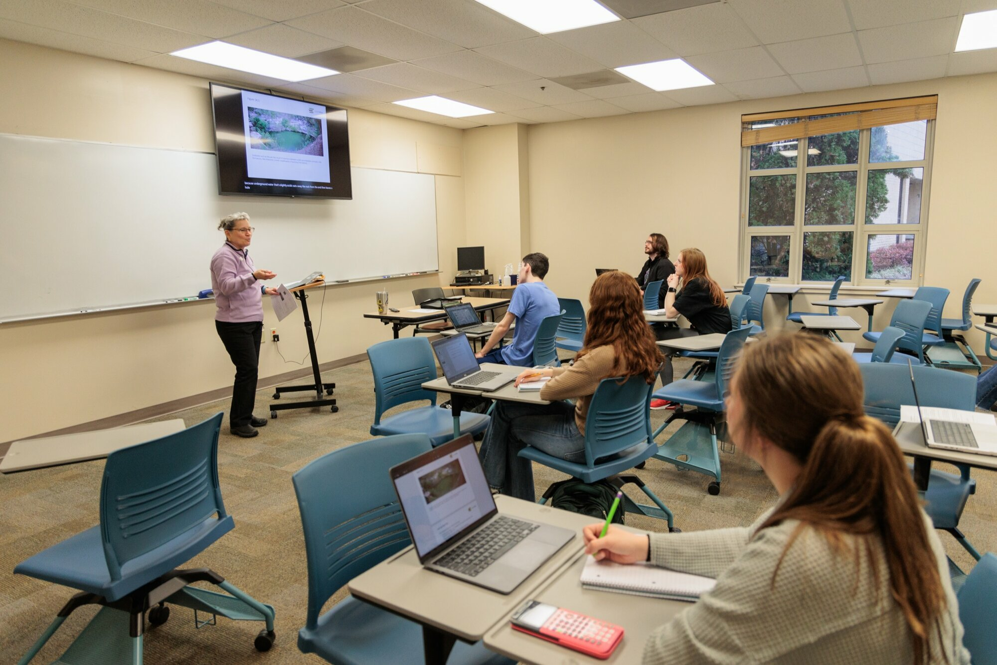 A college instructor presents at the front of a classroom, gesturing toward a screen displaying an image and text. Several students sit at desks with laptops and notebooks, listening attentively. The classroom features blue chairs, a large whiteboard, and natural light coming through the windows.