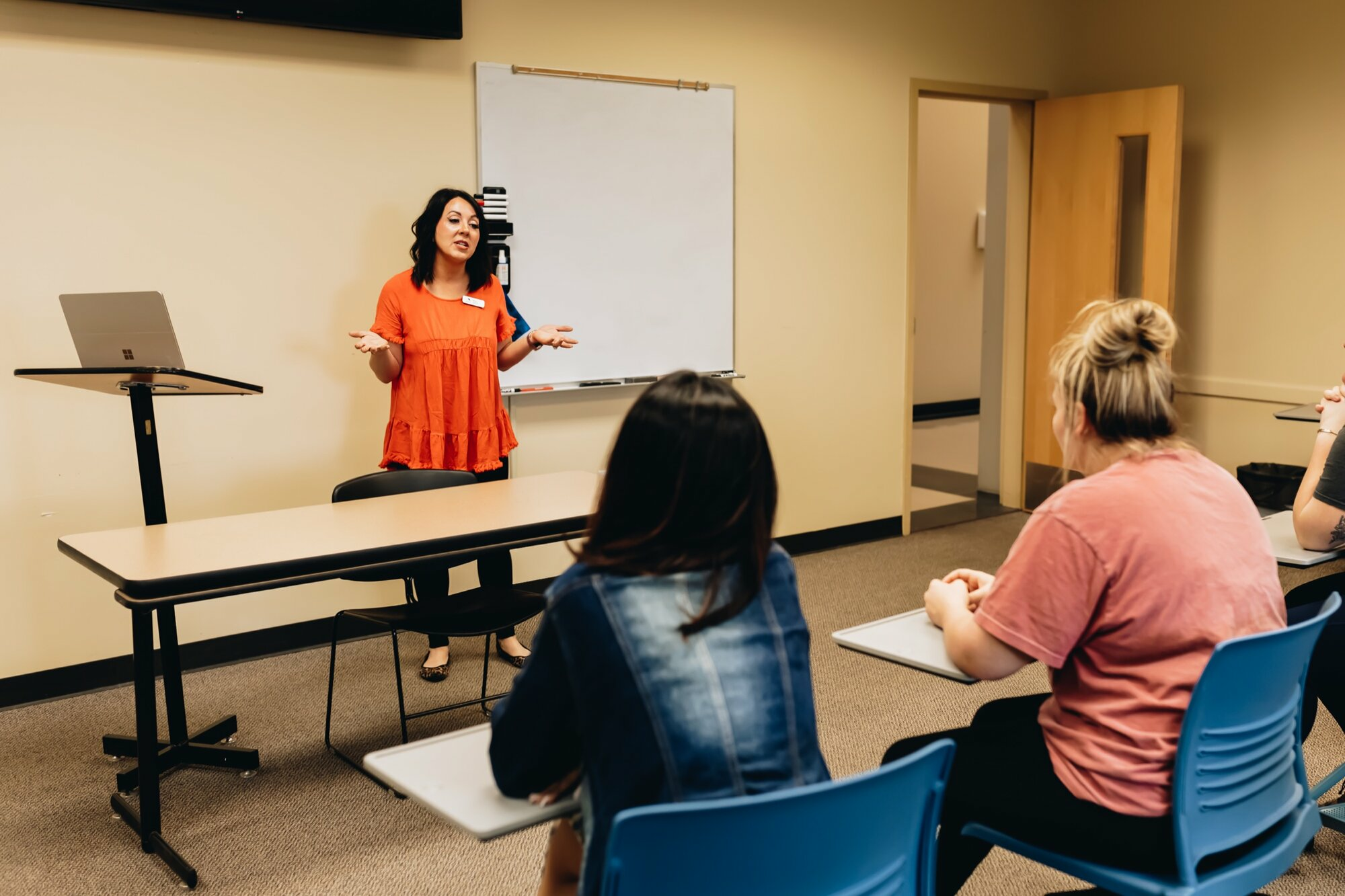 A college instructor stands at the front of a classroom speaking to students. She wears an orange blouse and gestures while addressing the class. Three students sit at desks facing her, listening attentively. A whiteboard and a laptop on a podium are visible in the background.