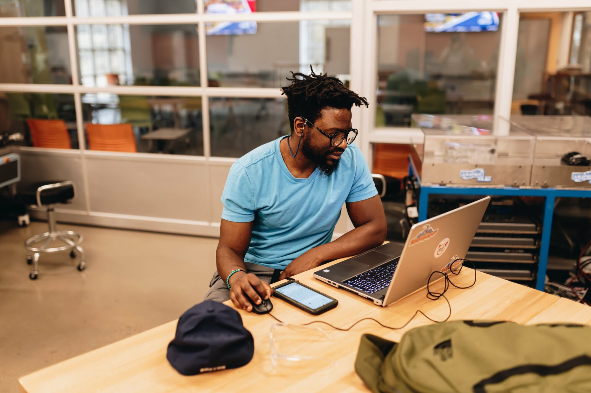 A student works on a laptop at a wooden table in a modern classroom or lab. He wears a light blue shirt and glasses, with earbuds in, focused on the screen. A phone, cap, backpack, and safety glasses rest on the table. Equipment and orange chairs are visible in the background through glass walls.