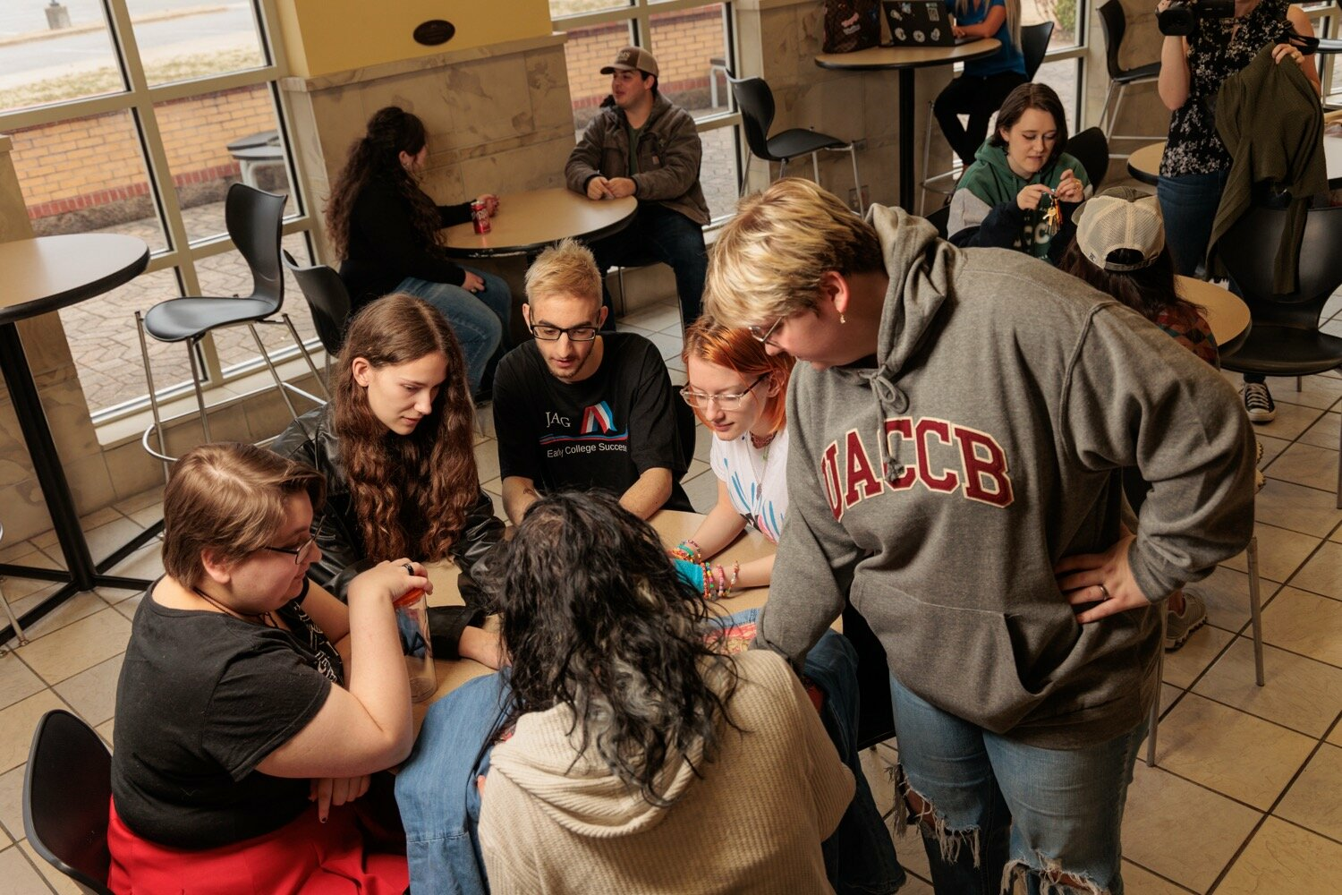 A group of UACCB students sit and stand around a table in the student commons, collaborating and talking while other students socialize in the background.