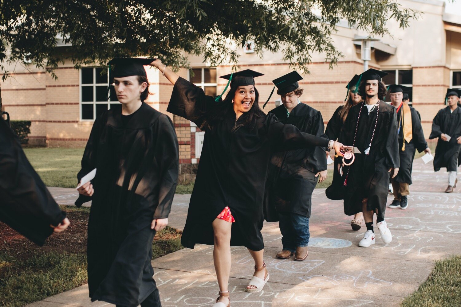 UACCB graduates walk in a campus procession wearing caps and gowns, with one graduate joyfully raising her arms and smiling as the group heads toward the ceremony.