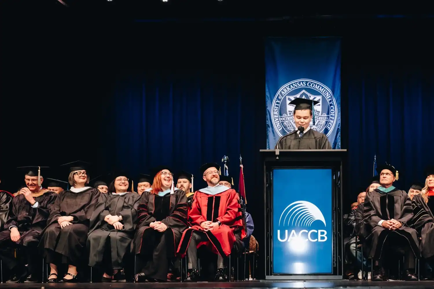 Zachary Heinecke, SGA President, in cap and gown stands at a podium delivering a commencement speech on a stage, with faculty in academic regalia seated behind them and a large UACCB banner and logo visible in the background.