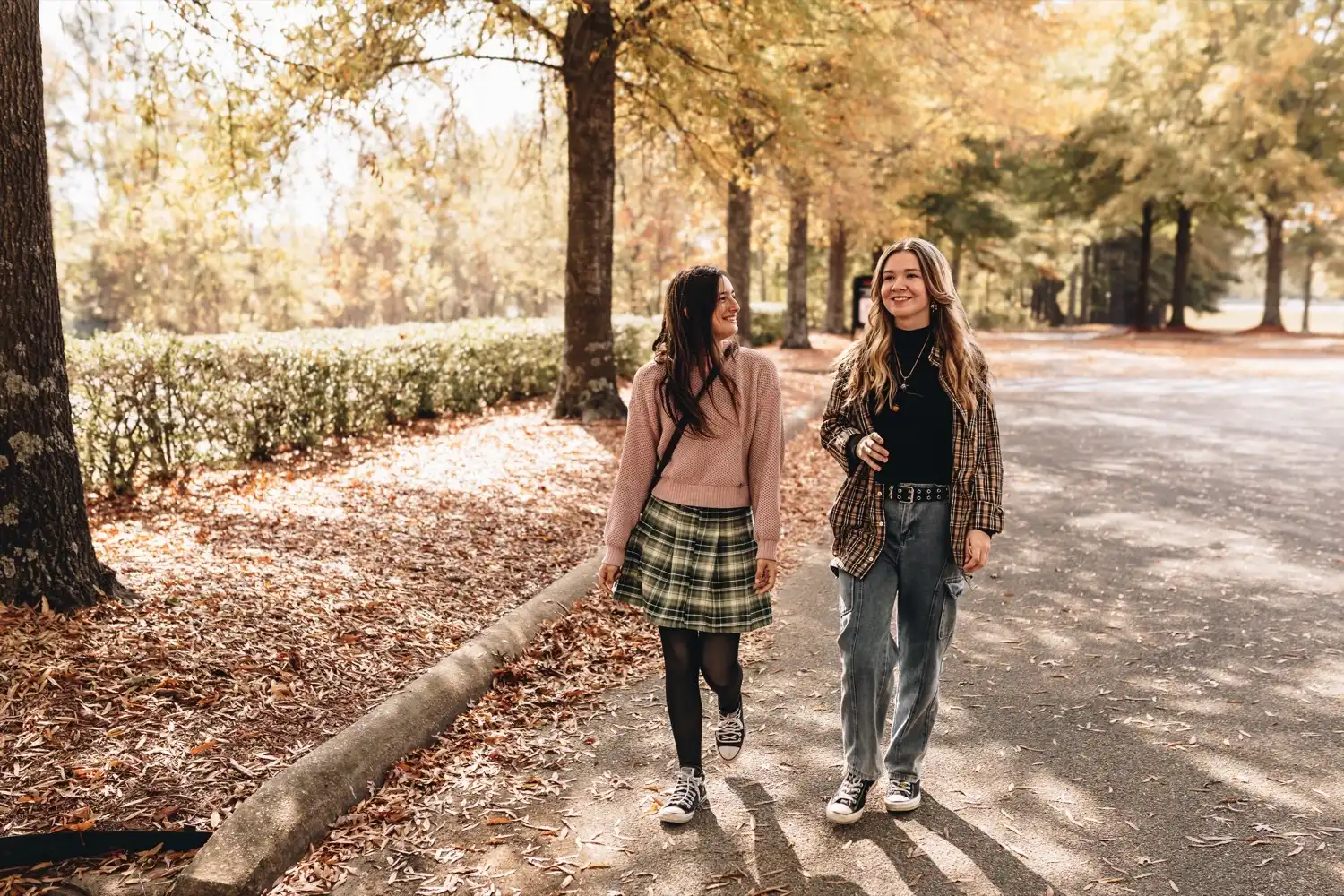 Two students walk and laugh together along a tree-lined campus path in autumn.