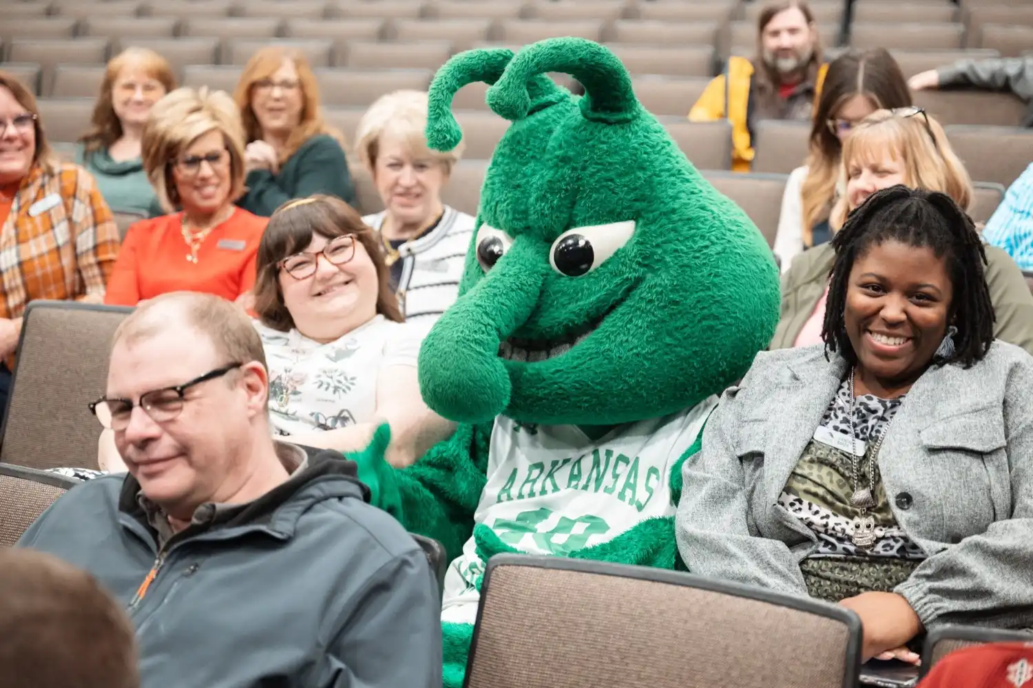 The University of Arkansas at Monticello mascot, Weezy, sits among smiling attendees in an auditorium during a partnership signing event.