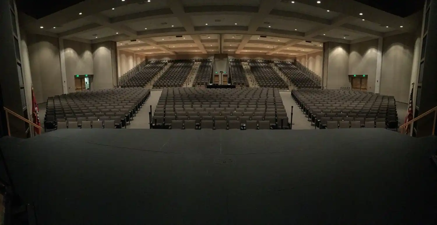 Interior of Independence Hall Auditorium viewed from the stage, showing 1,500 gray seats across a main floor and rear balcony beneath a coffered ceiling with recessed lighting.