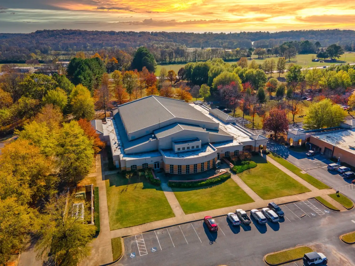 Aerial view of Independence Hall on the UACCB campus surrounded by fall foliage at sunset, with parking lots, green lawns, and tree-lined hills in the background