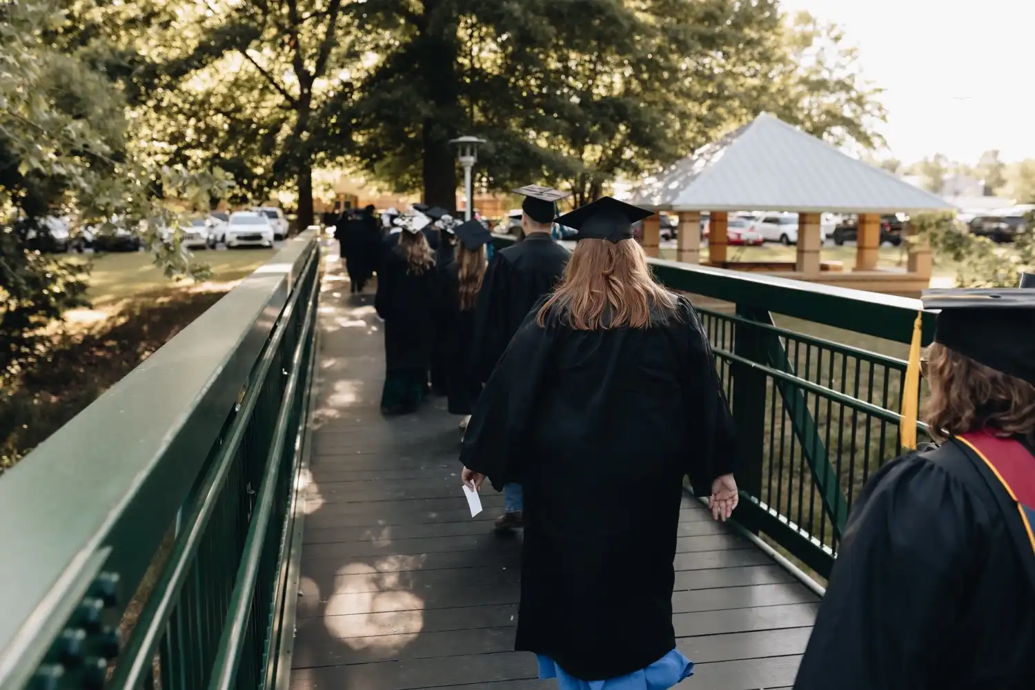 Graduates in caps and gowns walk across the campus walking bridge during UACCB graduation.