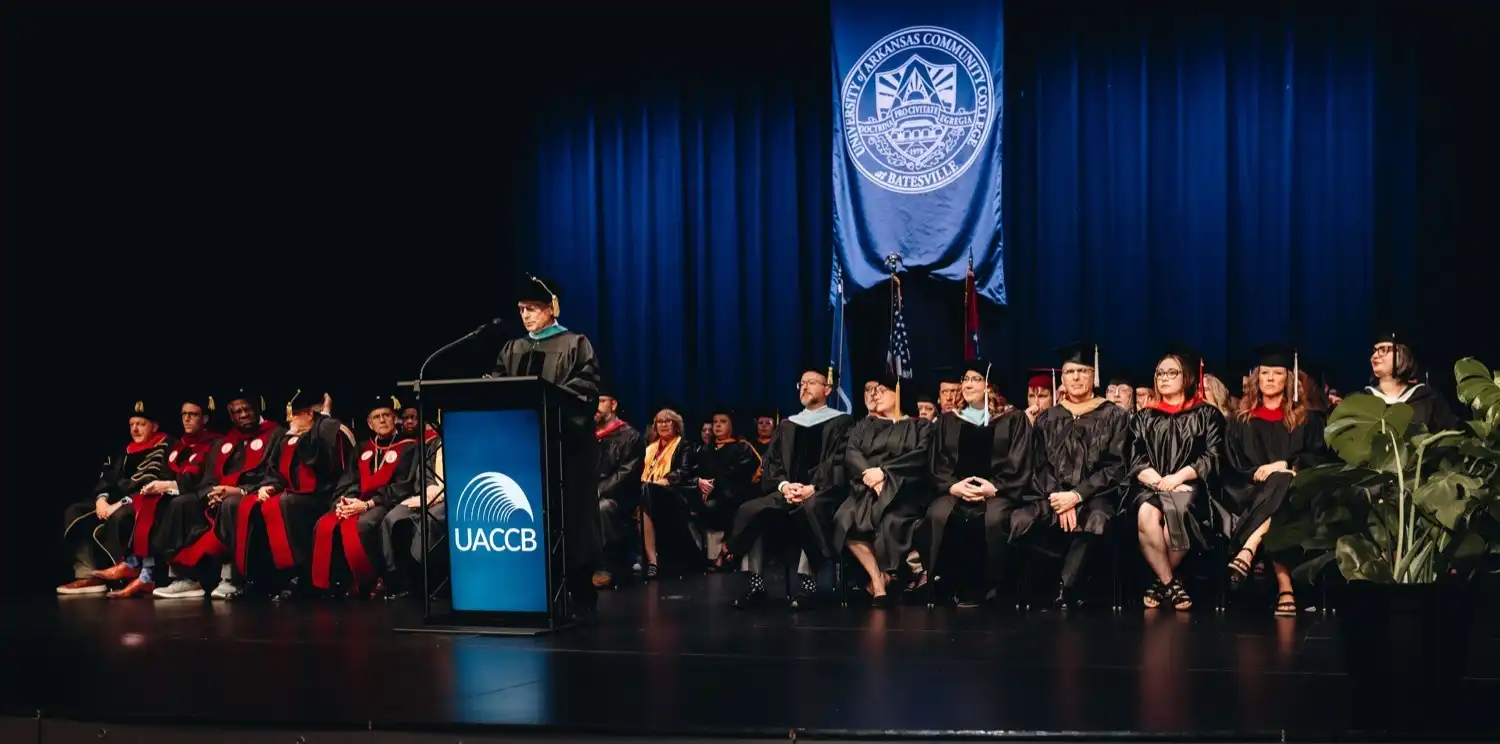 Dr. Brian Shonk, UACCB Chancellor, speaking at graduation with faculty and staff sitting on stage.