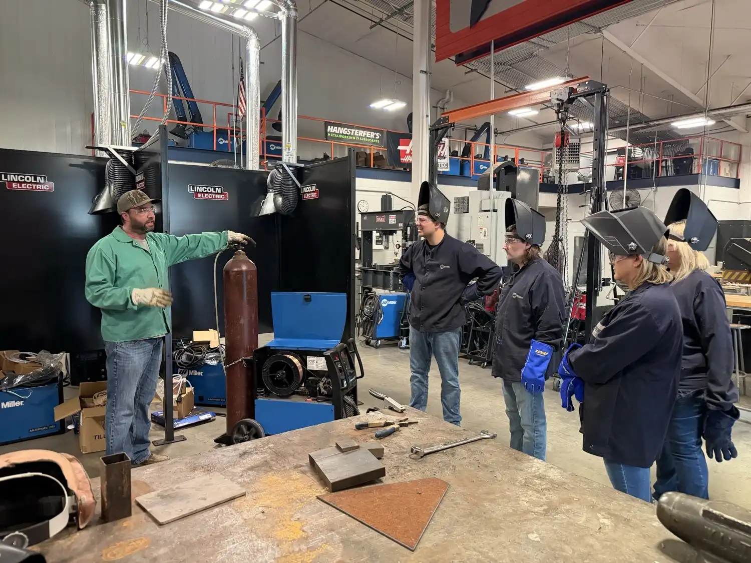 Justin Holman, welding instructor, speaks to UACCB Welcome Center employees wearing welding jackets, helmets, gloves, and safety glasses inside the welding lab.